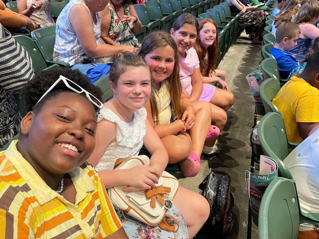 Group of students sitting in a row and smiling together for a selfie. The high school aged student in the front is holding the camera for the selfie.