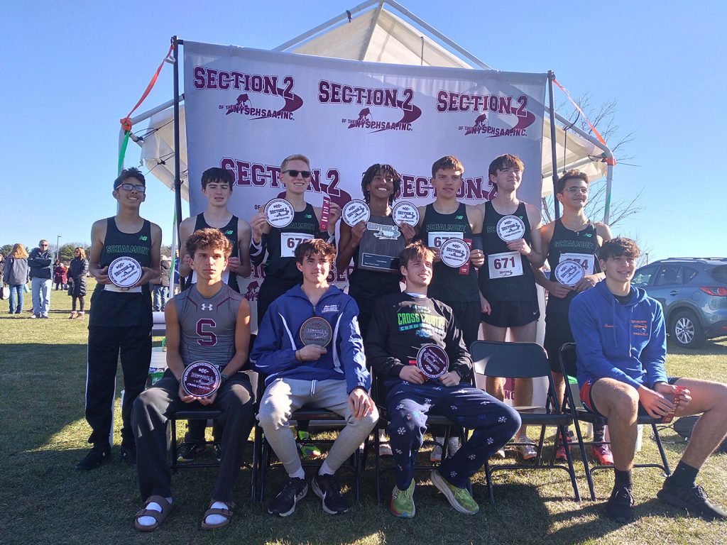 Boys cross country team holding individual awards and smiling for a photo. Four athletes are seated in front while several others are standing behind them.