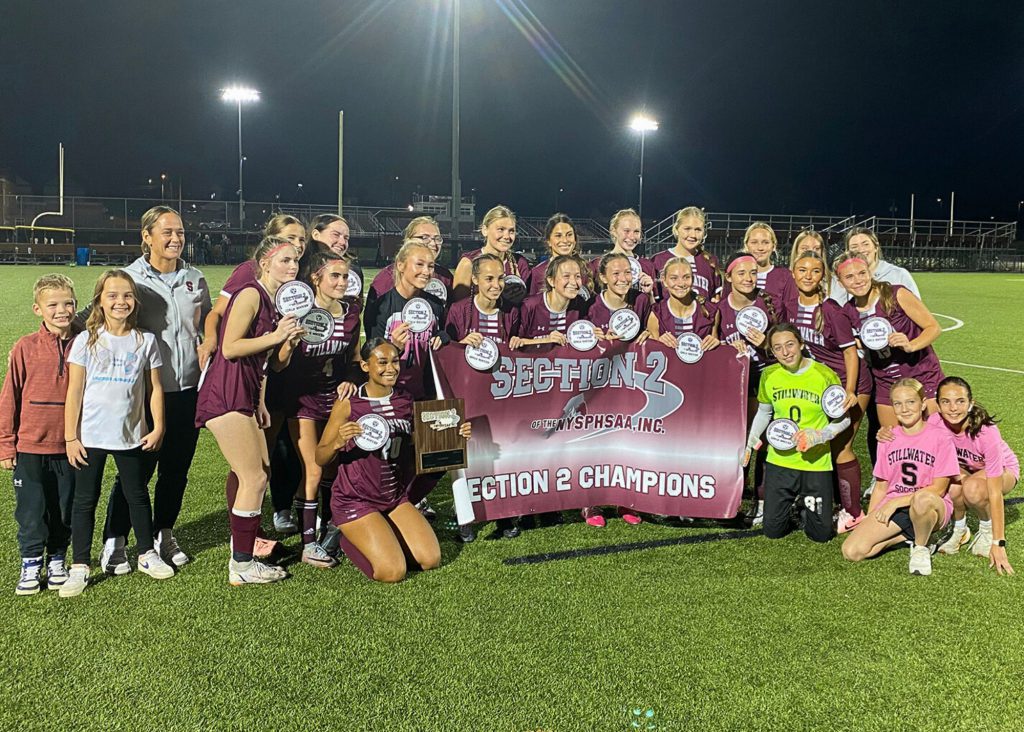 The girls varsity soccer team standing with their coach and her two children on the turf field, smiling for a group photo. The team is holding a banner that says "Section 2 Champions" and players are holding individual awards.