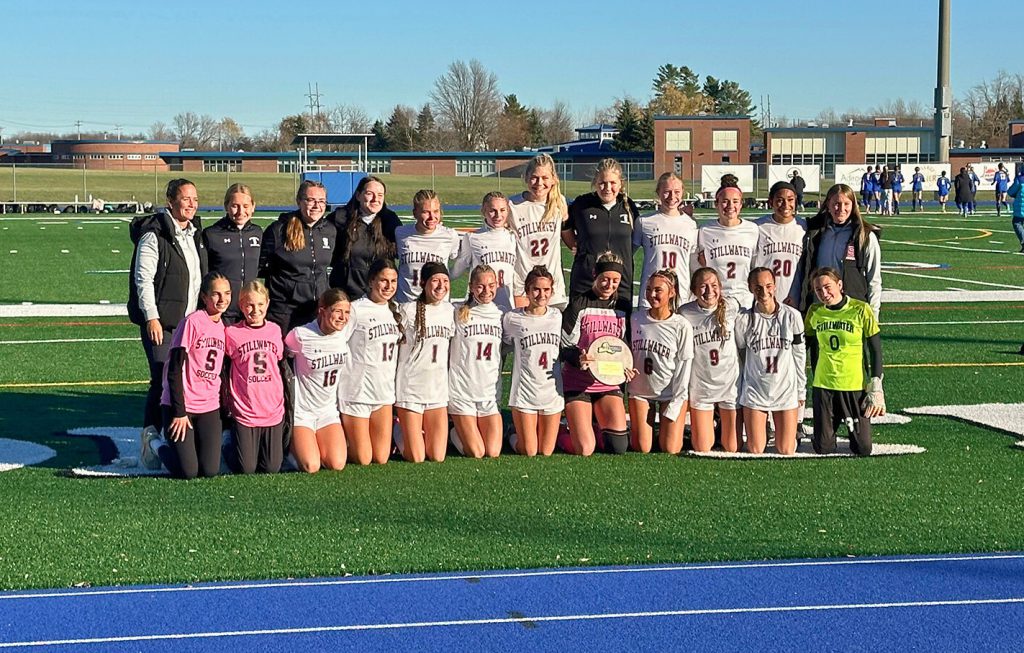 Girls varsity soccer team standing on the turf field together with their coach, smiling for a group photo. The goalie in the front row is holding an award.