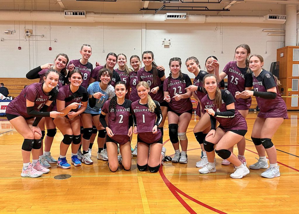 The girls varsity volleyball team standing together for a group photo. Two players are kneeling in front holding their shirts while the rest of the team members point to them. 