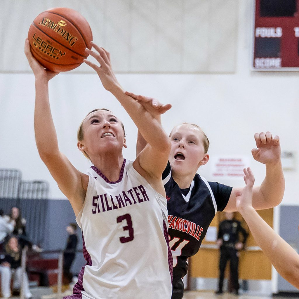 A girl in the maroon and white uniform attempts to shoot a lay up while another player in a black and red uniform attempts to block her shot.